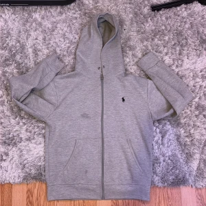 Grå hoodie från Polo Ralph Lauren - Snygg grå hoodie från Polo Ralph Lauren med dragkedja framtill och klassisk svart logga på bröstet. Tröjan har huva, två fickor och är tillverkad i mjuk bomull. Perfekt för en chill och stilren look. Har två mörka fläckar på framsidan, osäker om dem tvättas ut elr inte. Mvh