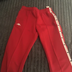 Röda mjukisbyxor med knappar - Röda mjukisbyxor  med vita sidor och svarta loggor längs benen. Byxorna har elastisk midja och tryckknappar längs sidorna för en sportig vibe. Perfekta för chill eller träning, med klassisk Kappa-stil.