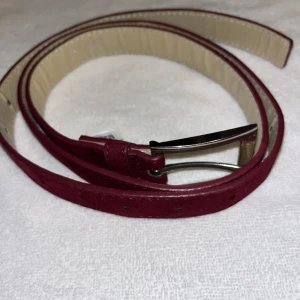 Mockabälte - Snyggt vinrött bälte i mocka med smal design och silverfärgat spänne. Insidan är beige och bältet har flera hål för justering. Perfekt accessoar för att lyfta din outfit med en stilren touch. Inga synliga defekter 