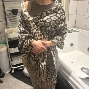 Leopardmönstrad beige halsduk med fransar - Stor och mysig halsduk med leopardmönster i beige, svart och bruna toner. Tjockt och mjukt material, perfekt att svepa runt sig. Fransar längs kanterna ger en extra touch. Snygg accessoar som sticker ut och håller dig varm. Kan använda på båda sidor olika färger. Ulliknade🤍🫶🏻