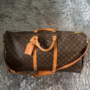 Louis Vuitton Keepall Monogram Resväska - Louis Vuitton Keepall i klassiskt monogrammönster med bruna och beige färger. Väskan har både handtag och axelrem i läder, guldiga metalldetaljer och dragkedja. Perfekt för resor med sin rymliga insida och ikoniska design.