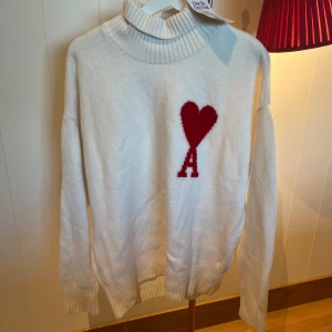Vit polotröja från AMI med rött hjärta - Vit polotröja från AMI med ribbad krage och muddar. På bröstet finns ett stort rött hjärta med bokstaven A. Tröjan är stickad och har lång ärm, perfekt för kyliga dagar. Snygg och trendig detalj som sticker ut.