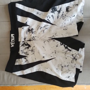 Venum svartvita träningsshorts - Grymma träningsshorts från Venum i svart och vitt med coolt batikmönster och logga på benet. Shortsen har bred midja med kardborreknäppning och är tillverkade i lätt syntetmaterial som andas. Perfekta för dig som kör MMA, kampsport eller annan intensiv träning.