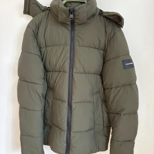 Olivgrön pufferjacka från Calvin Klein - Säljer en olivgrön dunjacka från Calvin Klein med skön huva och hög krage. Jackan har dragkedja framtill och logga på ärmen. Två fickor framgick innerficka med dragkedja. Bara använd ett fåtal gånger. Nypris 2900kr