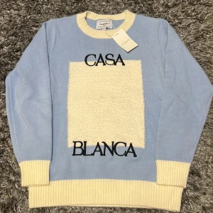 Ljusblå stickad tröja Casablanca M - Superfin ljusblå stickad tröja från Casablanca med krämvita ribbade muddar och krage. Stor vit, fluffig ruta framtill med svart broderad text 'CASA BLANCA'. Tillverkad i 100% bomull, perfekt för dig som gillar stilrena och trendiga plagg.