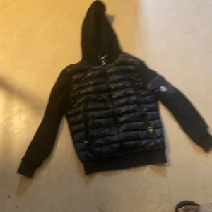 Svart Moncler  Cardigan dunjacka med huva - Svart dunjacka från Moncler med quiltad front och två dragkedjeförsedda fickor. Jackan har huva med snörning och Moncler-logga på ärmen. Ärmar och huva är i mjukt tyg medan kroppen är glansigt dunmaterial. Perfekt för kyliga dagar.