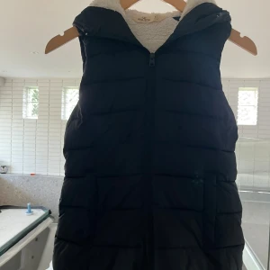 Svart dunväst med huva från Hollister - Svart väst från Hollister med mysigt vitt teddyfoder och huva. Västen har dragkedja framtill och två fickor på sidorna. Perfekt för lager-på-lager och chill höst- eller vinterstil.