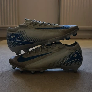 Nike Air Zoom fotbollsskor blå/vit - Säljer ett par Nike Air Zoom fotbollsskor i vitt med blå detaljer och snörning. Skorna har en smal, sportig siluett och är designade med dobbar för bästa grepp på planen. Tillverkade i syntetmaterial för lätt känsla och snabbhet. Perfekta för dig som vill dominera på fotbollsplanen.storlek 40