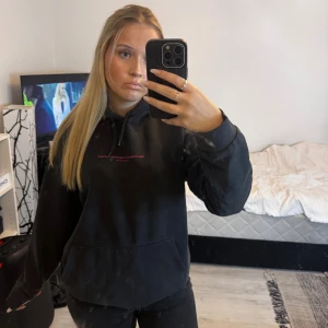 Svart hoodie med rosa graffiti-tryck - Säljer en svart hoodie med stor rosa graffiti-tryck på ryggen och mindre text på bröstet. Tröjan har huva, känguruficka och långa ärmar. Perfekt för dig som gillar streetstyle och vill sticka ut med ett statement-plagg.