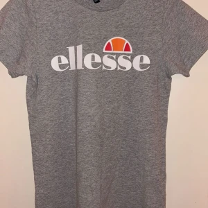 Grå t-shirt från Ellesse - Snygg grå t-shirt från Ellesse med klassisk logga i vitt, orange och rött framtill. T-shirten har rund halsringning och korta ärmar. Perfekt för en sportig och avslappnad stil.