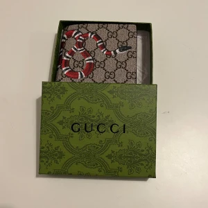 Plånbok med ormmotiv - Plånbok i brunt skinn med klassiskt GG-mönster och en röd, vit och svart orm på framsidan. Plånboken har flera kortfack och fönster för ID. Kommer i original grön box med mönster. Perfekt för dig som vill sticka ut med en lyxig accessoar.