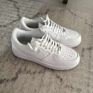 Nike Air Force 1 helvita sneakers - Klassiska Nike Air Force 1 sneakers i helvitt skinn med perforerad tå, vit sula och snörning. Ikonisk siluett med låg profil och diskret Nike-logga på sidan. Perfekta för dig som vill ha en clean och tidlös look.