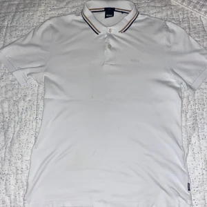 Vit pikétröja från BOSS - Vit pikétröja från BOSS i slim fit med klassisk krage och två knappar. Kragen har snygga ränder i marinblått och beige för en stilren touch. Tröjan är kortärmad och tillverkad i mjuk bomull, perfekt för en clean och fräsch look.