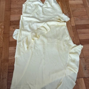 Ljusgul långklänning med volanger från H&M - Säljer en ljusgul långklänning från H&M i storlek S. Klänningen har v-ringad hals, ärmlös design och snygga volangdetaljer längs sidan och nederkanten. Materialet är mjukt och har en lätt glansig yta som ger en fräsch känsla. Perfekt för dig som gillar en feminin och somrig stil! Använd en gång 