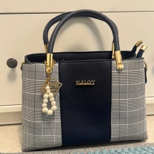 Rutig handväska från KLOY med pärldetalj -  Handväska i mörkblått skinnimitation med rutiga sidor i grått och vitt. Väskan har guldfärgade detaljer, pärlberlock och flera fack med dragkedja. 