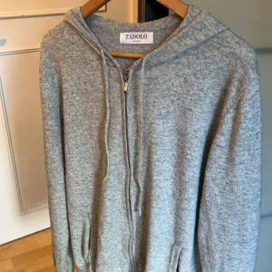 Grå Kashmir hoodie - Mjuk och stilren grå hoodie från Zadolo med hel dragkedja framtill, huva med snörning och två fickor. Tröjan är i ett stickat material som känns både bekvämt och snyggt. Perfekt för lager-på-lager och chill dagar.