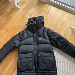Svart dunjacka Fjällräven Expedition M - Svart Fjällräven Expedition Series dunjacka med huva och gula snören. Jackan har två stora fickor framtill med knappar och dragkedja, samt klassisk Fjällräven-logga på bröstet. Perfekt för kalla dagar och riktigt snygg streetstil.