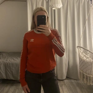 Röd Adidas tröja med vita ränder - Säljer en röd långärmad Adidas tröja med klassiska vita ränder längs ärmarna och broderad logga på bröstet. Tröjan har vit ribbad krage och är i mjuk bomull. Perfekt för en sportig och avslappnad stil.
