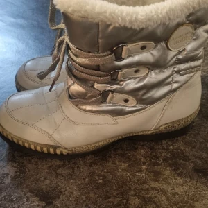 Silvriga vinterboots med snörning - Silvriga och vita vinterboots från Hogotex med fluffigt foder och glittriga snören. Skorna har quiltad ovandel, rund tå och platt sula. Perfekta för kalla dagar med sin varma insida och snygga metallic-look. En liten smutsfläck på fodret på ena sidan. Se bild.