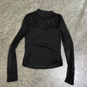Svart mesh polotopp med mönster - Svart långärmad polotopp i mesh med coolt geometriskt mönster upptill och på ärmarna. Toppen har en hög hals och är figurnära med transparenta partier som ger en edgy vibe. Perfekt att styla lager på lager eller som statement-piece.