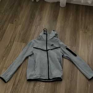 Grå Nike zip-hoodie med svarta detaljer - Snygg grå hoodiejacka från Nike med hel dragkedja, svarta detaljer och logga på bröstet. Jackan har huva, lång ärm och en cool svart detalj på ena ärmen. Perfekt för en sportig och avslappnad stil.