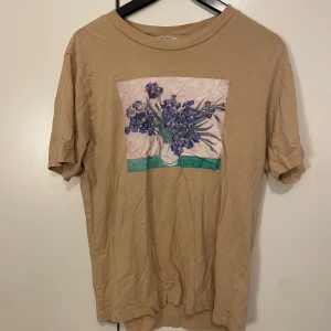 Beige t-shirt med blommigt tryck - Beige t-shirt med ett stort tryck av lila blommor i vas på bröstet. T-shirten har rund hals och korta ärmar, perfekt för en chill och konstnärlig vibe. Materialet känns mjukt och skönt mot huden.