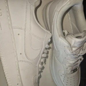Nike Air Force 1  - Klassiska Nike Air Force 1 sneakers i helvitt skinn med vit sula och vita snören. Ikonisk design med perforerad tå, broderad Nike-logga på hälen och metallbricka med 'AF1' på snörningen. Perfekta för dig som gillar en clean och tidlös look.