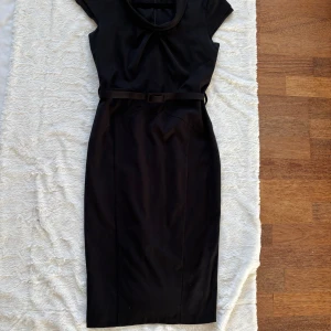 Svart midiklänning från Warehouse - Stilren svart klänning från Warehouse med snygg krage, korta ärmar och bälte i midjan. Klänningen har en figurnära passform och dragkedja i ryggen. Perfekt för dig som gillar klassisk och tidlös stil.