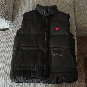 Svart dunväst från Canada Goose - Svart dunväst från Canada Goose med hög krage, stora framfickor och röd broderad lönnlöv på bröstet. Västen har knappar framtill och klassisk logga på ena fickan. Perfekt för kyliga dagar när du vill hålla stilen.