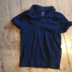 Mörkblå pikétröja från H&M - En stilren mörkblå pikétröja från H&M med klassisk krage och korta ärmar. Tröjan är tillverkad i mjuk bomull och har en enkel, tidlös design utan mönster eller tryck. Perfekt för en clean och avslappnad look.