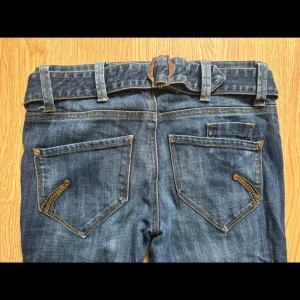 Vintage bootcut jeans - Snygga blå jeans med bootcut-modell och coolt bälte i midjan. Jeansen har klassiska fickor med dekorativa sömmar och detaljer i bronsfärg. Materialet är jeans med en lätt tvättad look och markerade sömmar. Perfekta för dig som gillar en avslappnad men trendig stil.