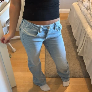 Ljusblå bootcut jeans med fickdetaljer - Säljer ett par ljusblå jeans med bootcut passform och låg midja. Jeansen har snygga fickor med lock och knapp bak samt klassiska femficksdetaljer fram. Tillverkade i mjukt denimtyg som är jätte sköna. 