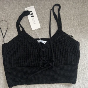 Svart stickad linnetopp FB Sister - Svart stickad linnetopp från FB Sister med smala axelband och snygg snörning framtill. Toppen har en croppad passform och är perfekt för att skapa en trendig look. Materialet är mjukt och följsamt stickat.