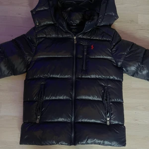 Svart pufferjacka från Polo Ralph Lauren - Svart pufferjacka från Polo Ralph Lauren med röd logga på bröstet. Jackan har huva, dragkedja framtill och tre fickor med dragkedja. Den är vadderad och har en glansig finish, perfekt för kalla dagar.