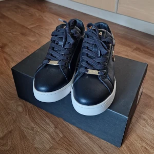 Svarta sneakers från Mexx med gulddetaljer - Snygga svarta sneakers från Mexx med vita platta sulor och gulddetaljer på snörningen och sidan. Skorna är i skinnimitation med blank häl och rund tå. Perfekta för dig som vill ha en stilren och trendig look. Använda 1 gång, liten i storlek.