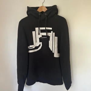 Svart hoodie från J.Lindeberg - Säljer en snygg svart hoodie från J.Lindeberg med ett coolt vitt grafiskt tryck på framsidan. Den har en klassisk passform med långa ärmar. Hoodien är använd men har mycket kvar att ge!
