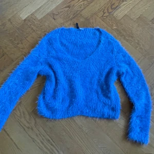 Blå fluffig stickad tröja Gina Tricot - Supermjuk och fluffig blå stickad tröja från Gina Tricot med v-ringning och lång ärm. Tröjan har en cool, lite lurvig textur som gör den extra mysig och trendig. Perfekt att styla med jeans eller kjol för en chill look.