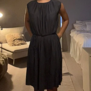 Svart plisserad kortklänning med knyt - Säljer en svart plisserad klänning med knyt i midjan. Klänningen är ärmlös och har en avslappnad passform med rund halsringning.