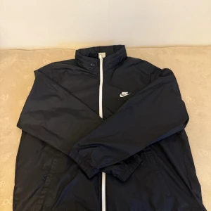 Svart tunn jacka från Nike - Svart vindjacka från Nike med vit dragkedja och klassisk logga på bröstet. Jackan har hög krage, två sidofickor och är tillverkad i lätt polyester. Perfekt för blåsiga dagar och enkel att matcha med streetwear.