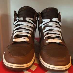 Nike Dunk High bruna sneakers - Nike Dunk High sneakers i brunt skinn med vita detaljer och vit swoosh på sidan. Skorna har högt skaft, vita snören och en brun yttersula. Klassisk siluett med rund tå och platt sula, perfekt för dig som vill ha en avslappnad men ändå snygg outfit