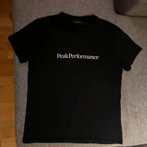 Svart Peak Performance t-shirt - Svart t-shirt från Peak Performance med vit logga tryckt på bröstet. Klassisk rund hals och korta ärmar. Tillverkad i mjuk bomull för en skön känsla och enkel stil.