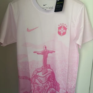 Nike Brasil fotbollströja rosa - Snygg fotbollströja från Nike i ljusrosa med tryck av Kristusstatyn och Rio de Janeiro. Bröstet har Nike-logga och Brasilien CBF-märke. Tillverkad i lätt, ventilerande Dri-FIT-material som passar perfekt för träning eller match.