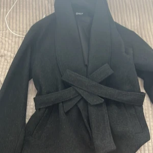 Svart kappa med bälte från ONLY - Stilren svart kappa från ONLY med bred krage och knytskärp i midjan. Jackan har en klassisk, rak passform och är perfekt för dig som vill ha en snygg och enkel look. Fodrad insida för extra komfort.