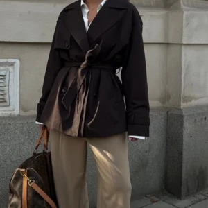 Brun trenchcoat med bälte - Snygg mörkbrun trenchcoat med klassisk krage, dubbelknäppning och bälte i midjan. Jackan har långa ärmar med spänndetaljer vid handlederna och en stilren, rak passform. Från NAKD, strl 38. Endast använd 1 gång