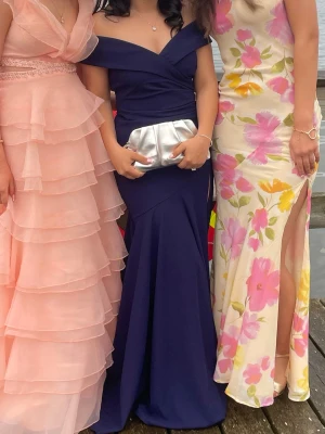 Marinblå långklänning offshoulder från Bubbleroom  - Snygg marinblå långklänning med offshoulder och v-ringad urringning. Klänningen har figurnära passform och en elegant slits framtill. Perfekt för dig som vill sticka ut med en klassisk och stilren look. Använd endast en gång. 