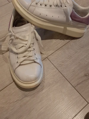 Alexander McQueen vita sneakers - Stilrena vita sneakers från Alexander McQueen med tjock sula och lila detalj på hälen. Klassisk låg modell med perforerade sidor och breda vita skosnören. Perfekta för dig som gillar en clean och trendig look.