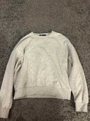 Ljusgrå sweatshirt från FB Sister - En basic ljusgrå sweatshirt från FB Sister med rund halsringning och ribbade muddar vid ärmslut och nederkant. Tröjan har en avslappnad passform och är tillverkad i mjukt sweatshirtmaterial, perfekt för en chill och enkel stil. Det är storlek M men kan verka mindre i storleken