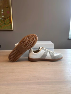 Skor från Masion margiela - Stilrena sneakers i vitt och beige med snygga mockadetaljer och bruna gummisulor. Skorna sälj på grund av att de inte passade på mig. Svarar gärna på alla frågor. Storlek 43.