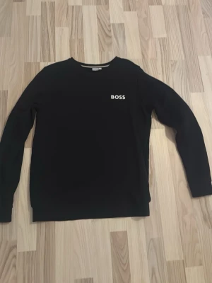 Svart tröja från BOSS i bomull - Svart långärmad tröja från BOSS med enkel och clean design. Liten vit BOSS-logga på bröstet. Skön passform och rund halsringning, perfekt till jeans eller chinos. Tillverkad i mjuk bomull för en bekväm känsla. Skriv gärna vid funderingar!