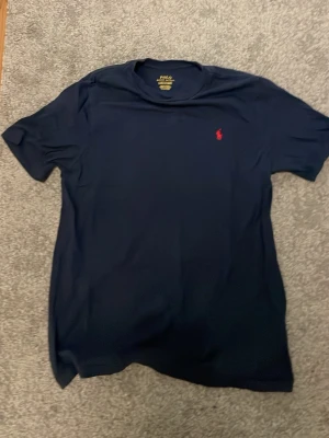 Mörkblå t-shirt från Polo Ralph Lauren - Snygg mörkblå t-shirt från Polo Ralph Lauren med klassisk röd logga broderad på bröstet. T-shirten har rund halsringning och korta ärmar, perfekt för en stilren och enkel look. Pris kan sänkas 
